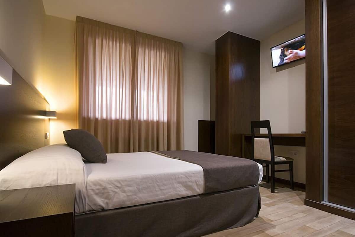 A hotel room with a bed, a desk, a chair, a TV mounted on the wall, and a window with curtains.