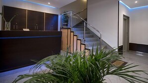 A modern reception area with a dark reception desk, a glass staircase, and a potted plant. - Complejo O Piñeiral (Fonsagrada)