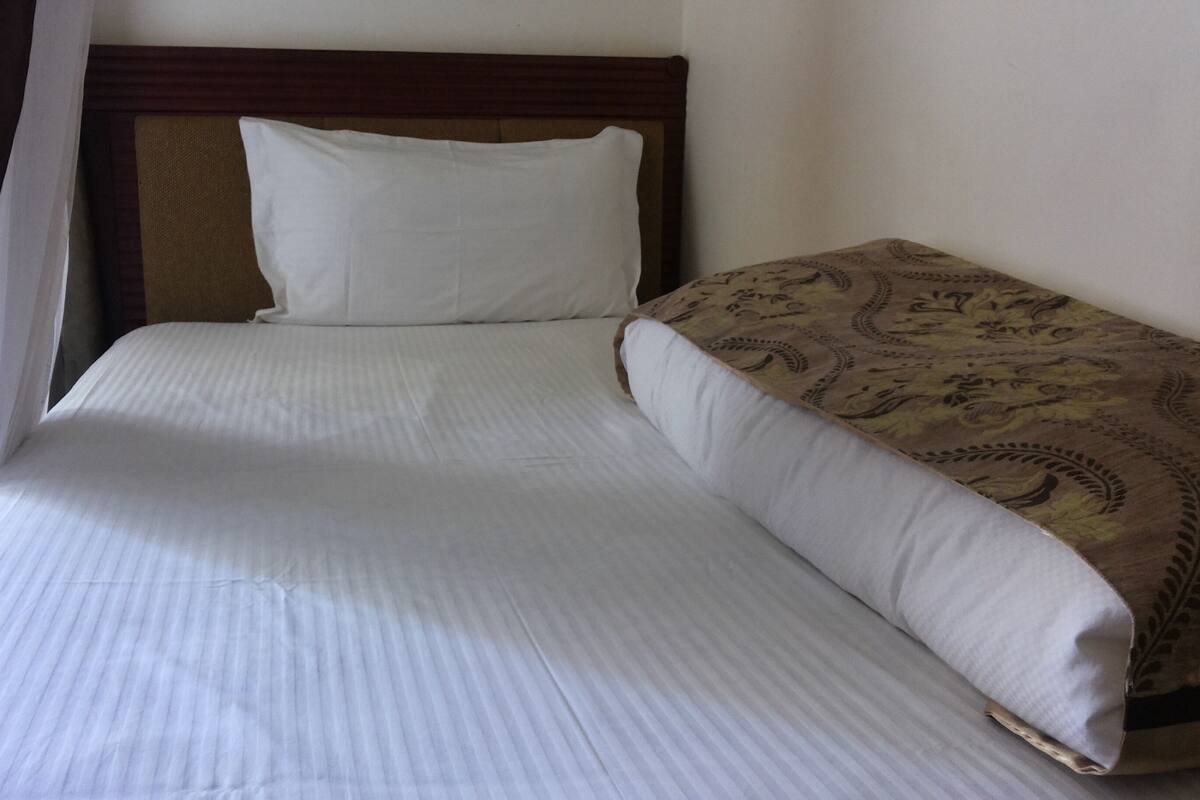 A single bed with a white mattress and a patterned blanket, a wooden headboard, and a plain wall.