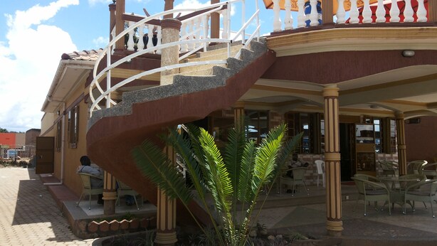 A building with a staircase leading to a balcony, outdoor seating area, and a small garden with a palm tree. - Hotel Bucolic Heritage (Adjumani)