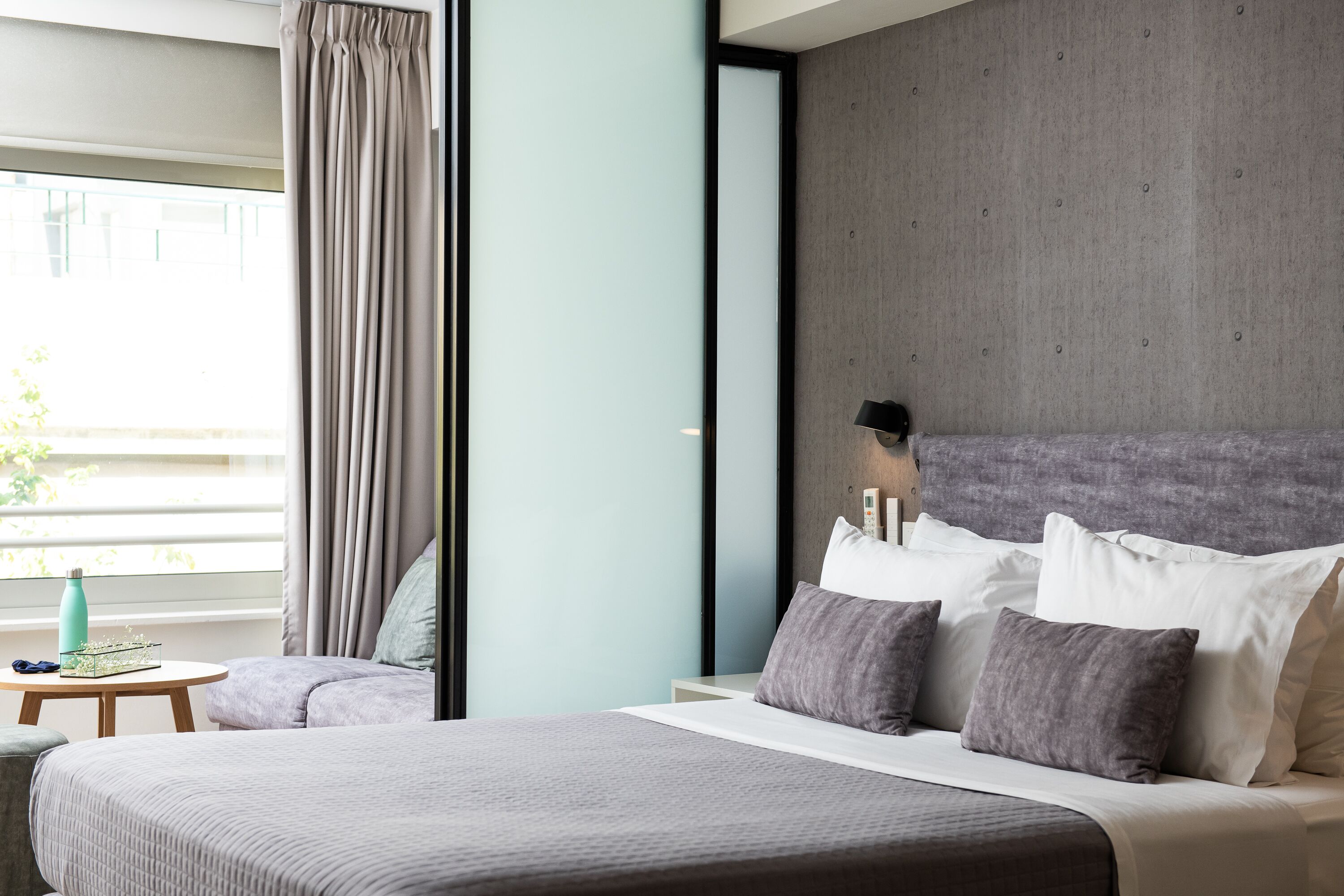 A modern hotel room with a large bed, a bedside table, a lamp, and a view of the outdoors.
