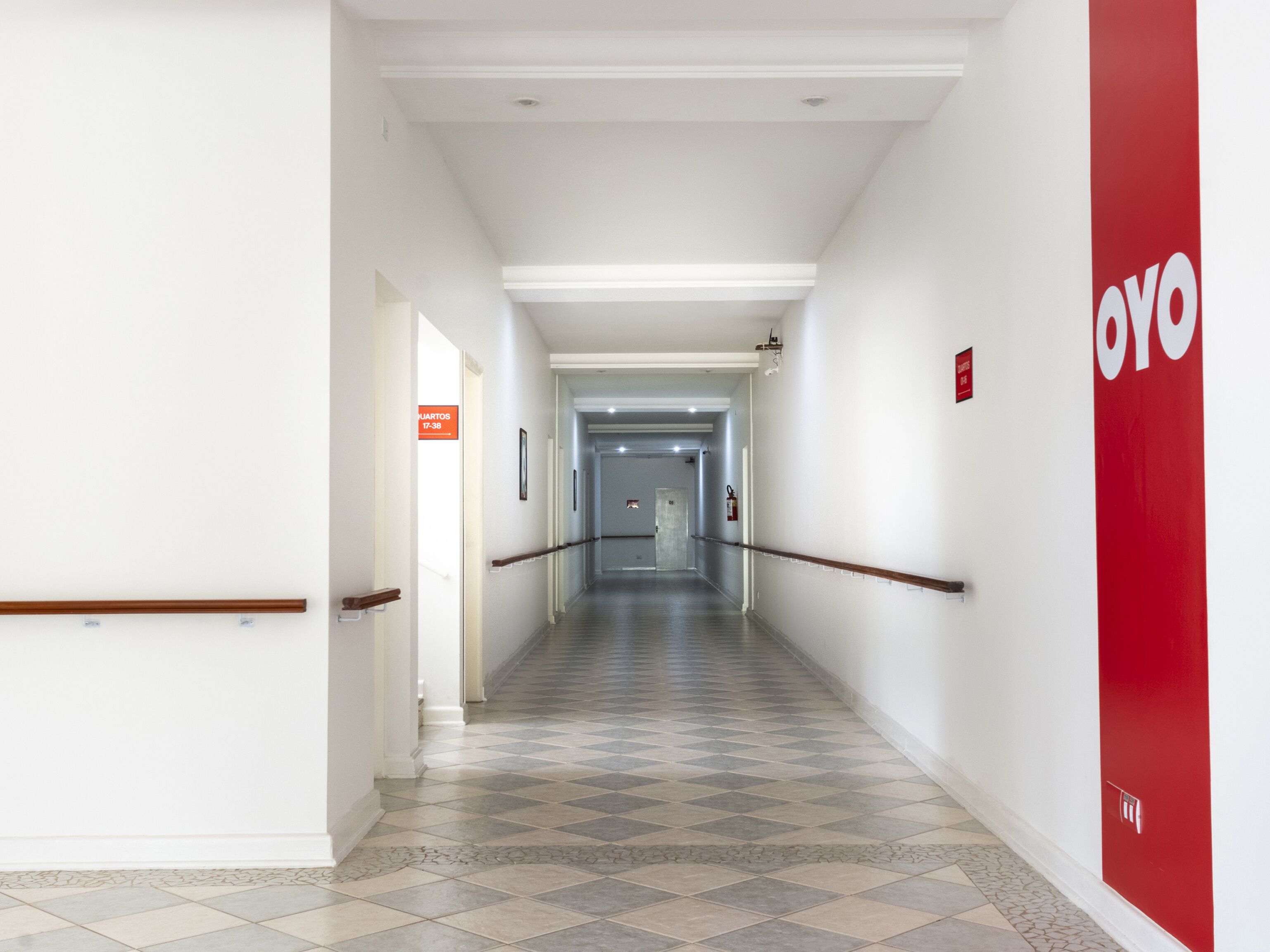 Um corredor de hotel com piso xadrez, paredes brancas e uma placa vermelha à direita.