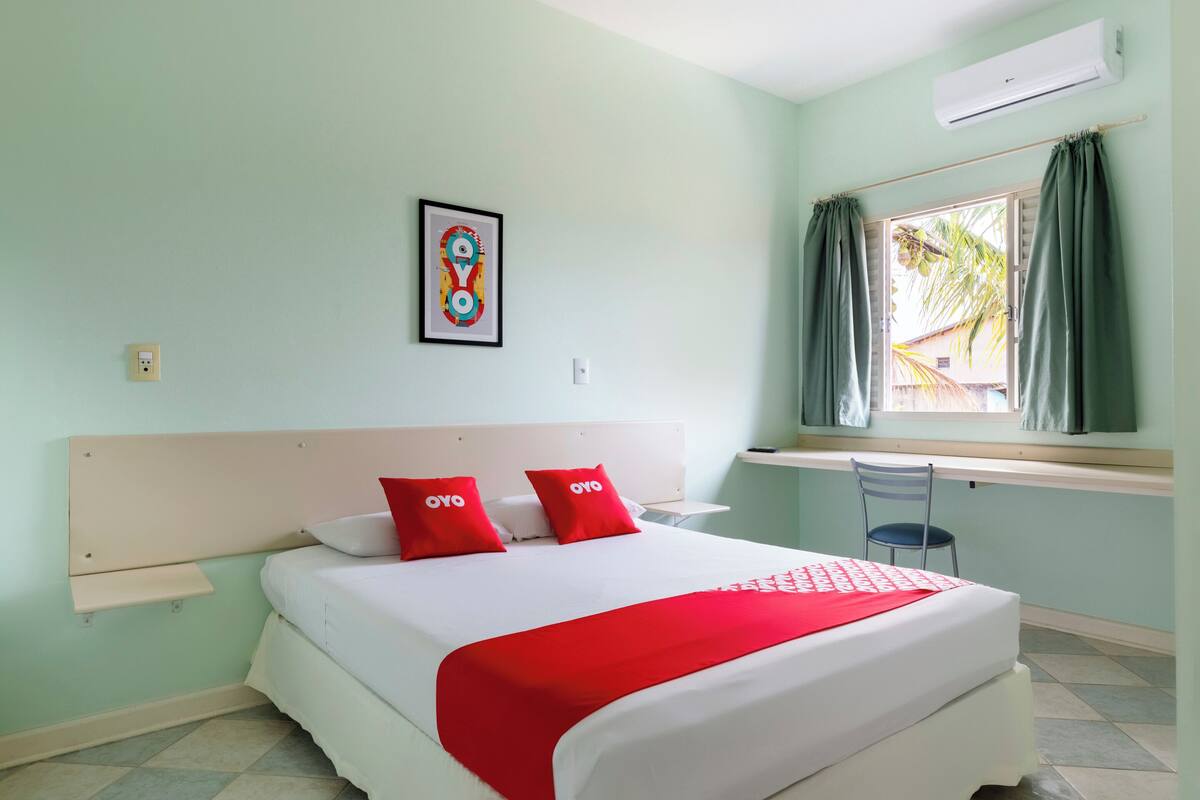 Una cama bien tendida con almohadas rojas, cabecera blanca y una manta roja. Hay una mesita con una silla junto a una ventana con cortinas.