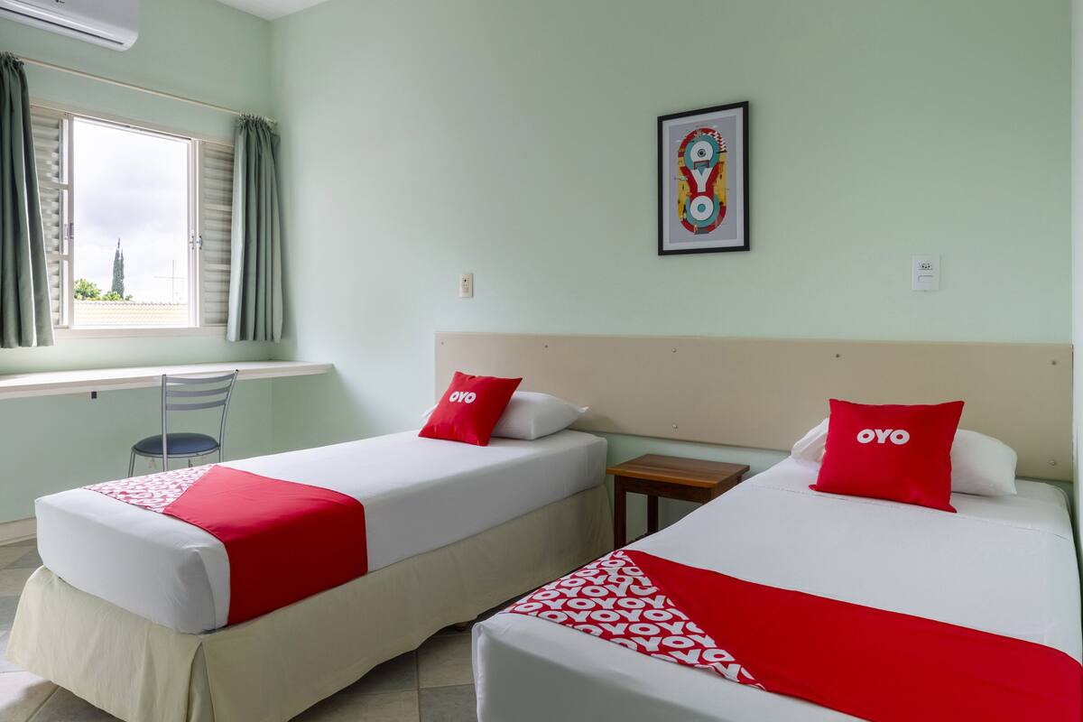 Habitación de hotel con dos camas, cada una con colcha y almohadas rojas y blancas, una mesita de madera, una silla y un ventanal con cortinas.