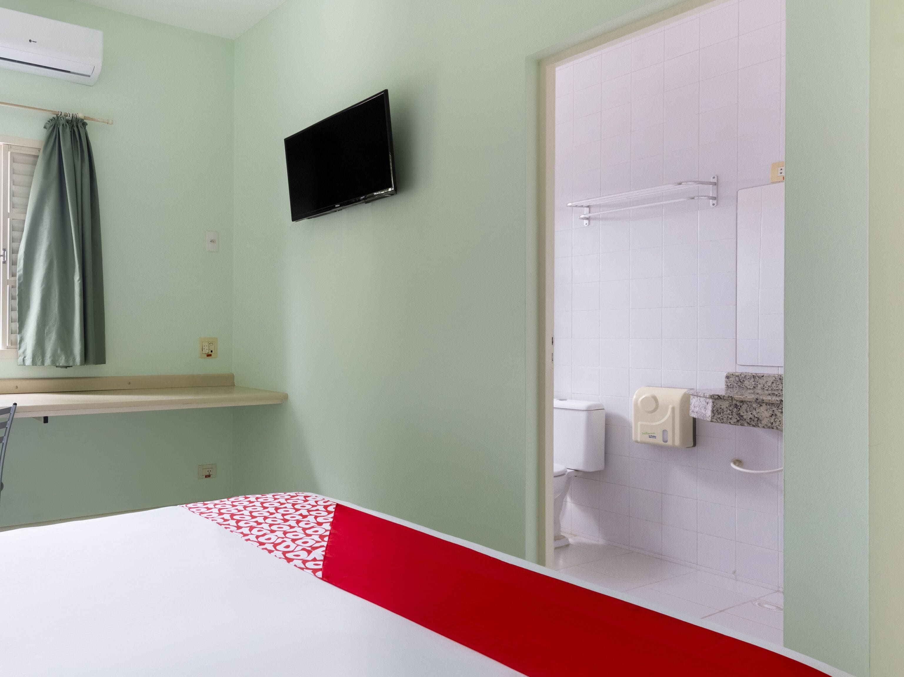 A hotel room with a bed, a television mounted on the wall, a window with curtains, a shower area with a towel rack, and a toilet.