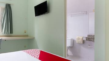 A hotel room with a bed, a television mounted on the wall, a window with curtains, a shower area with a towel rack, and a toilet.