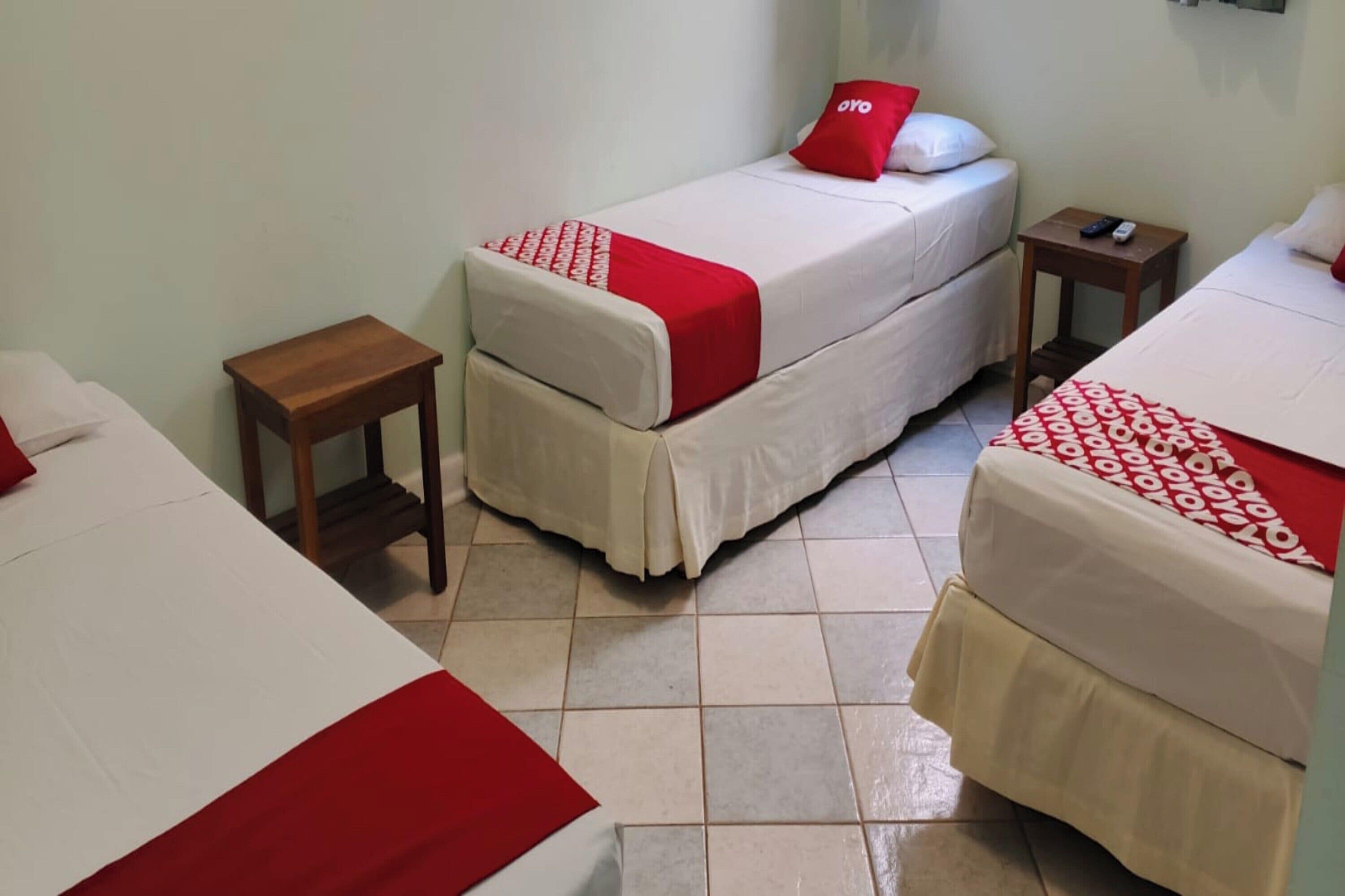 Habitación de hotel con dos camas individuales, cada una con cubrecama rojo y blanco, una mesita de noche de madera y un control remoto sobre la mesita.