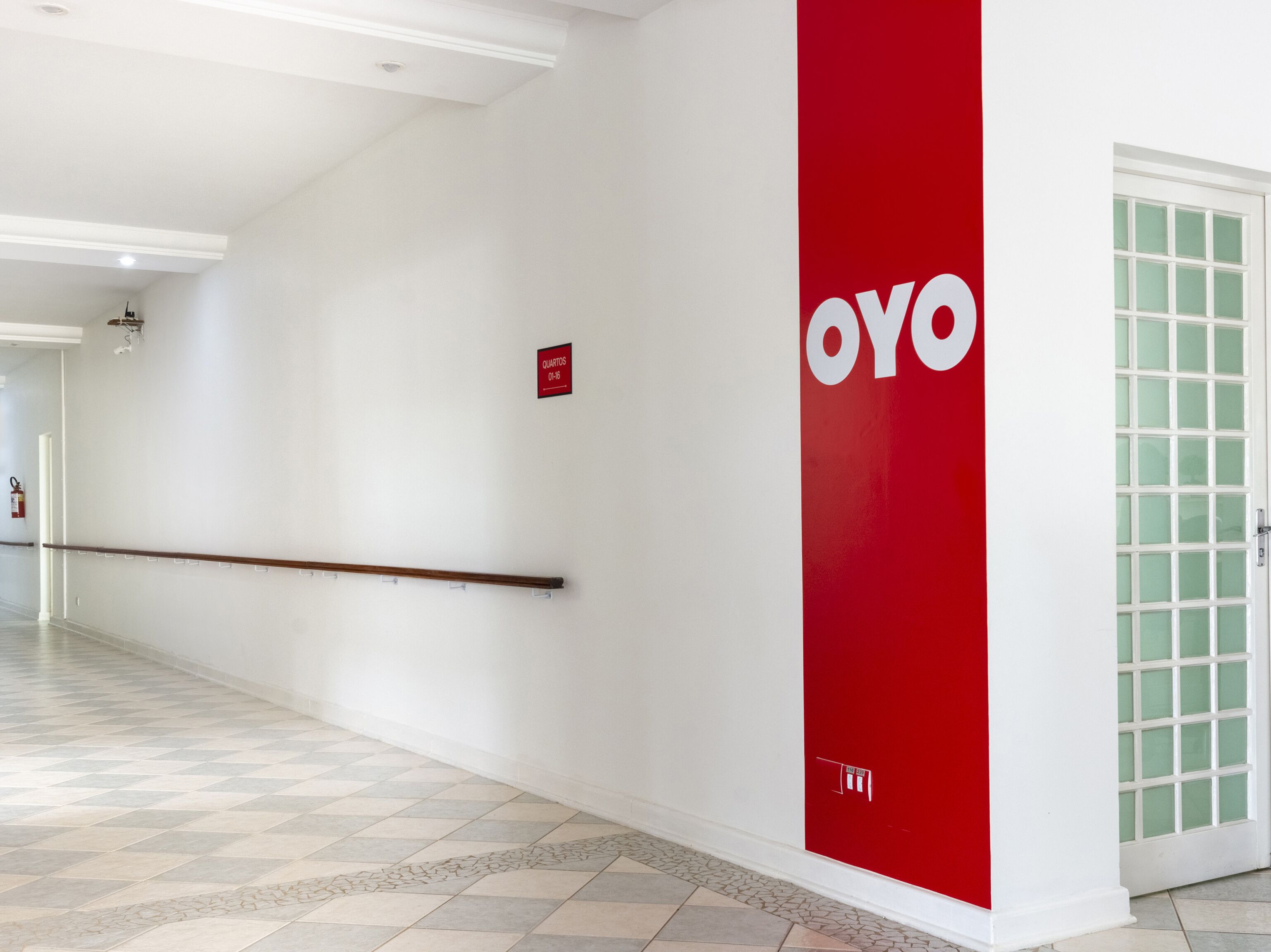 Un pasillo con una pared roja que muestra el logotipo 'OYO', un extintor y una puerta con paneles de vidrio satinado.