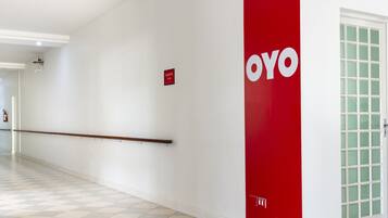 Un pasillo con una pared roja que muestra el logotipo 'OYO', un extintor y una puerta con paneles de vidrio satinado.