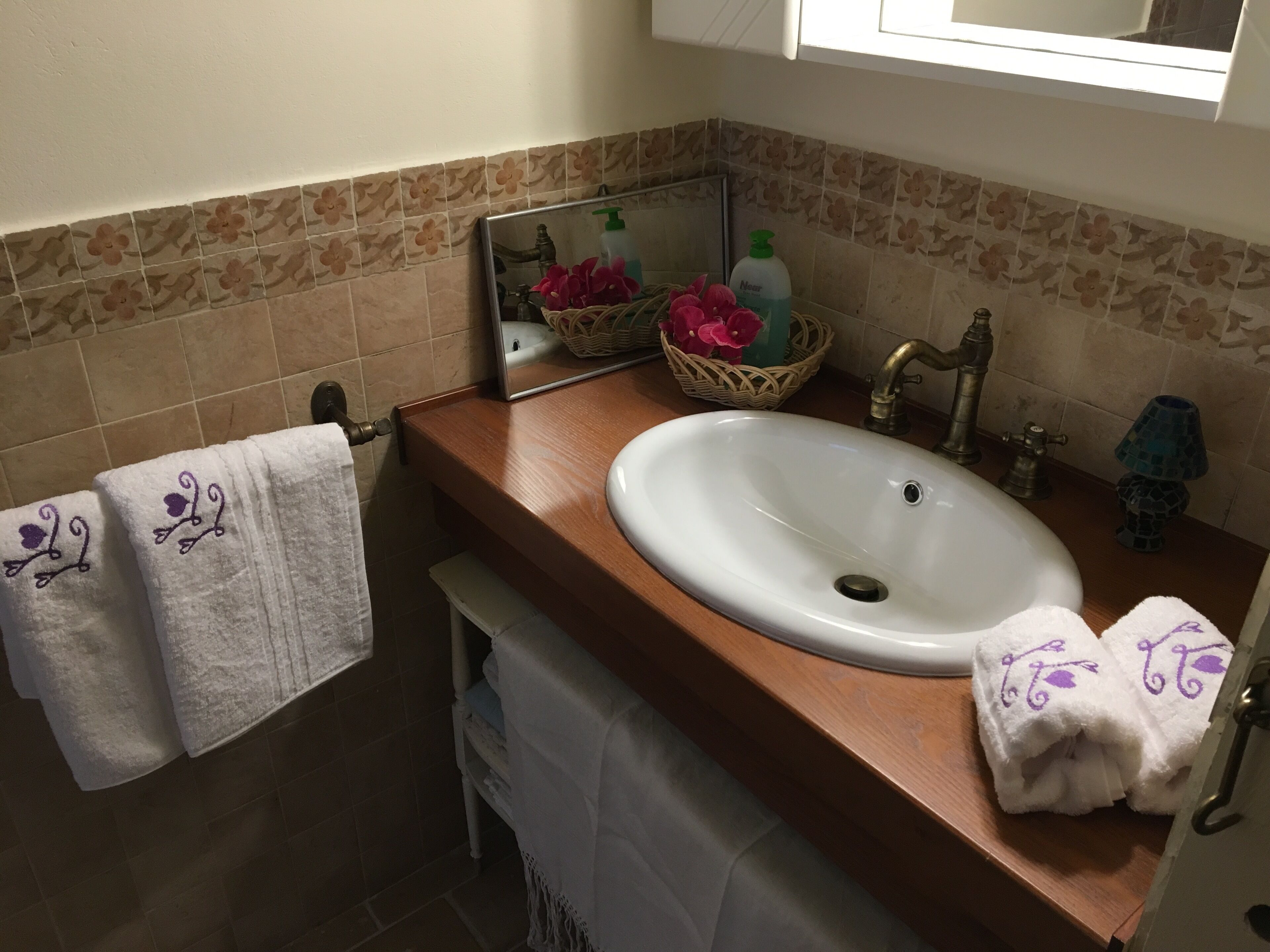 Un baño con un mueble de tocador de madera, un lavabo blanco, un espejo, un perchero con toallas, una cesta decorativa y una lámpara pequeña.