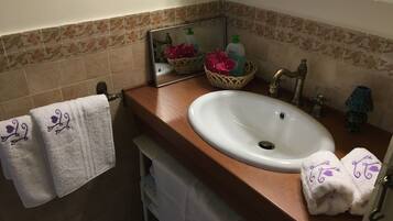 Un baño con un mueble de tocador de madera, un lavabo blanco, un espejo, un perchero con toallas, una cesta decorativa y una lámpara pequeña.