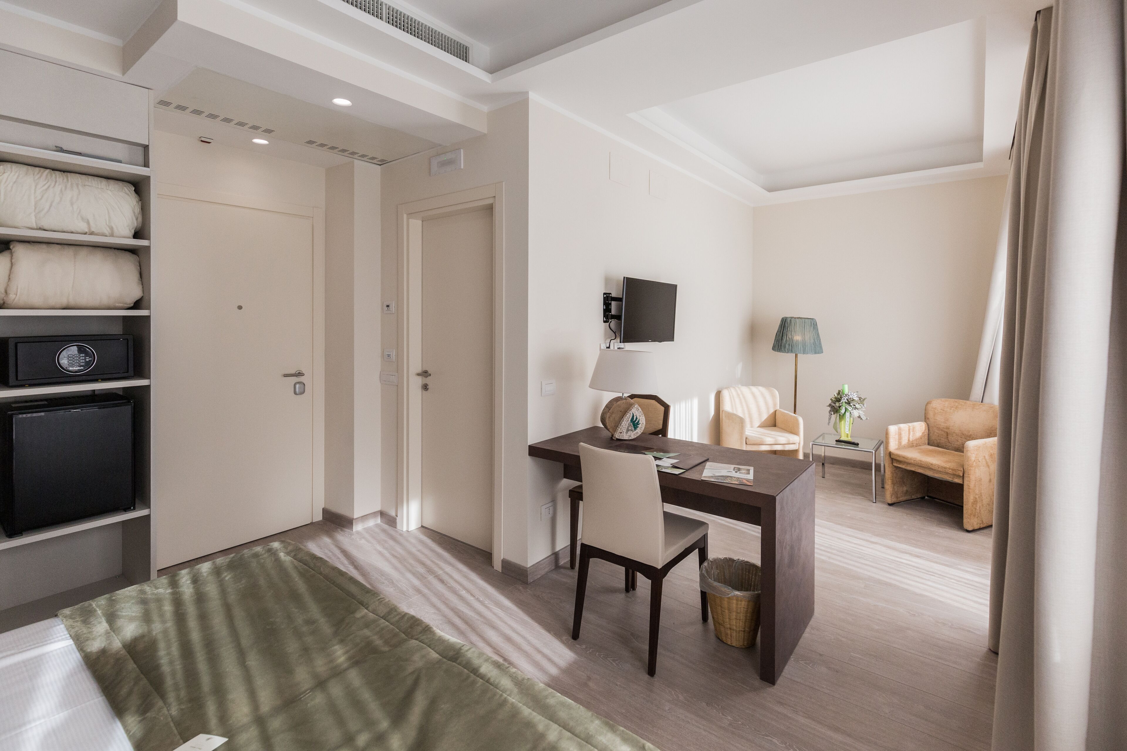 A hotel room with a desk, chair, TV, sofa, and a small table with a lamp.