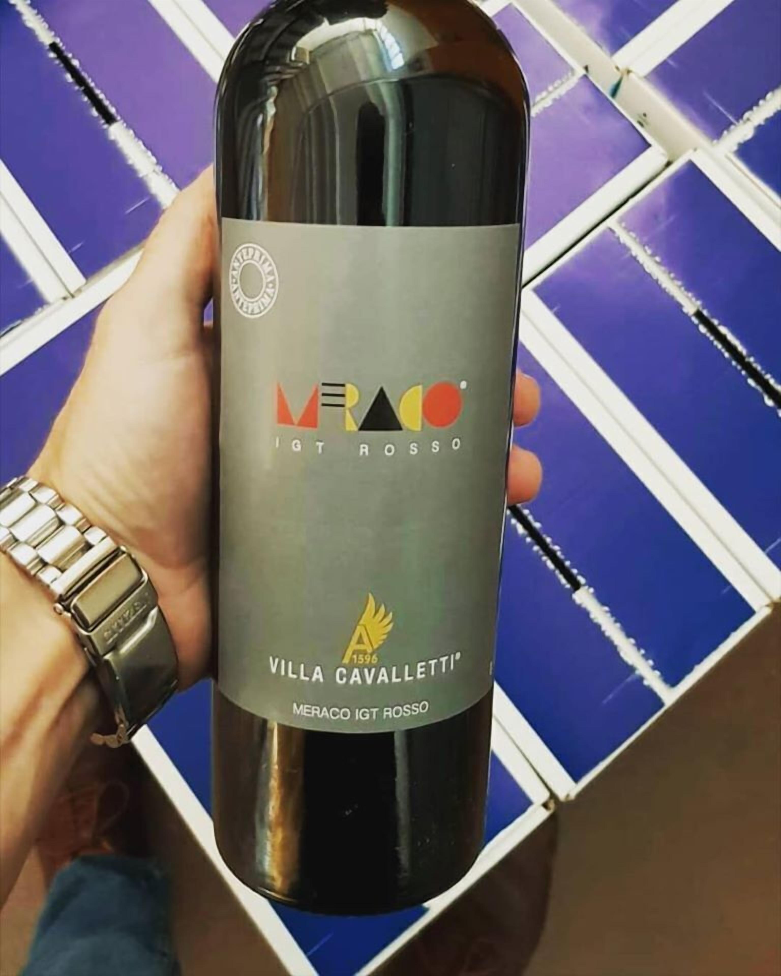 Een persoon houdt een fles Meraco IGT Rosso wijn vast, met een etiket waarop 'VILLA CAVALLETTI' staat.