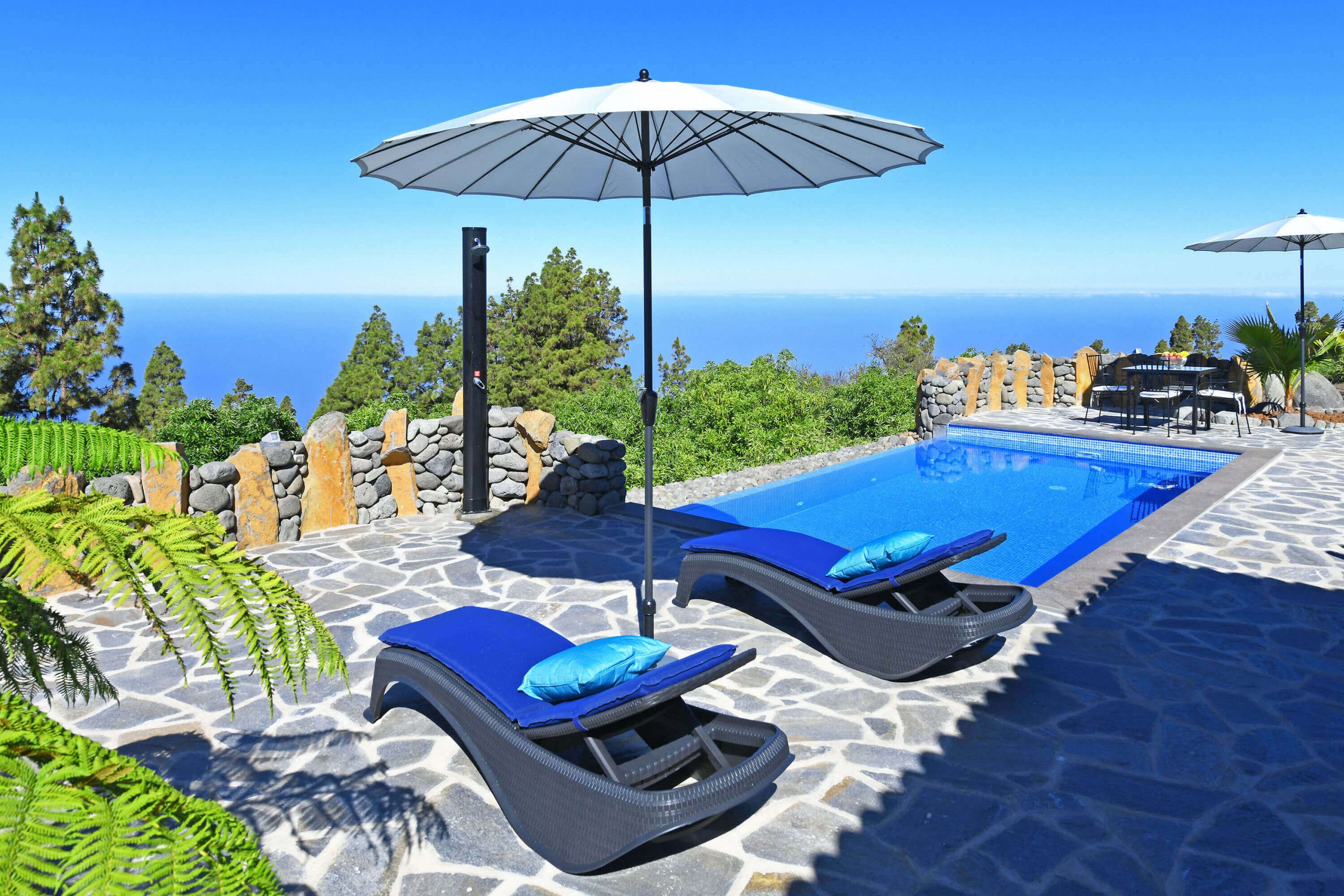Un área junto a la piscina con dos sillas de descanso y sombrillas, una terraza de baldosas de piedra y vista al océano.