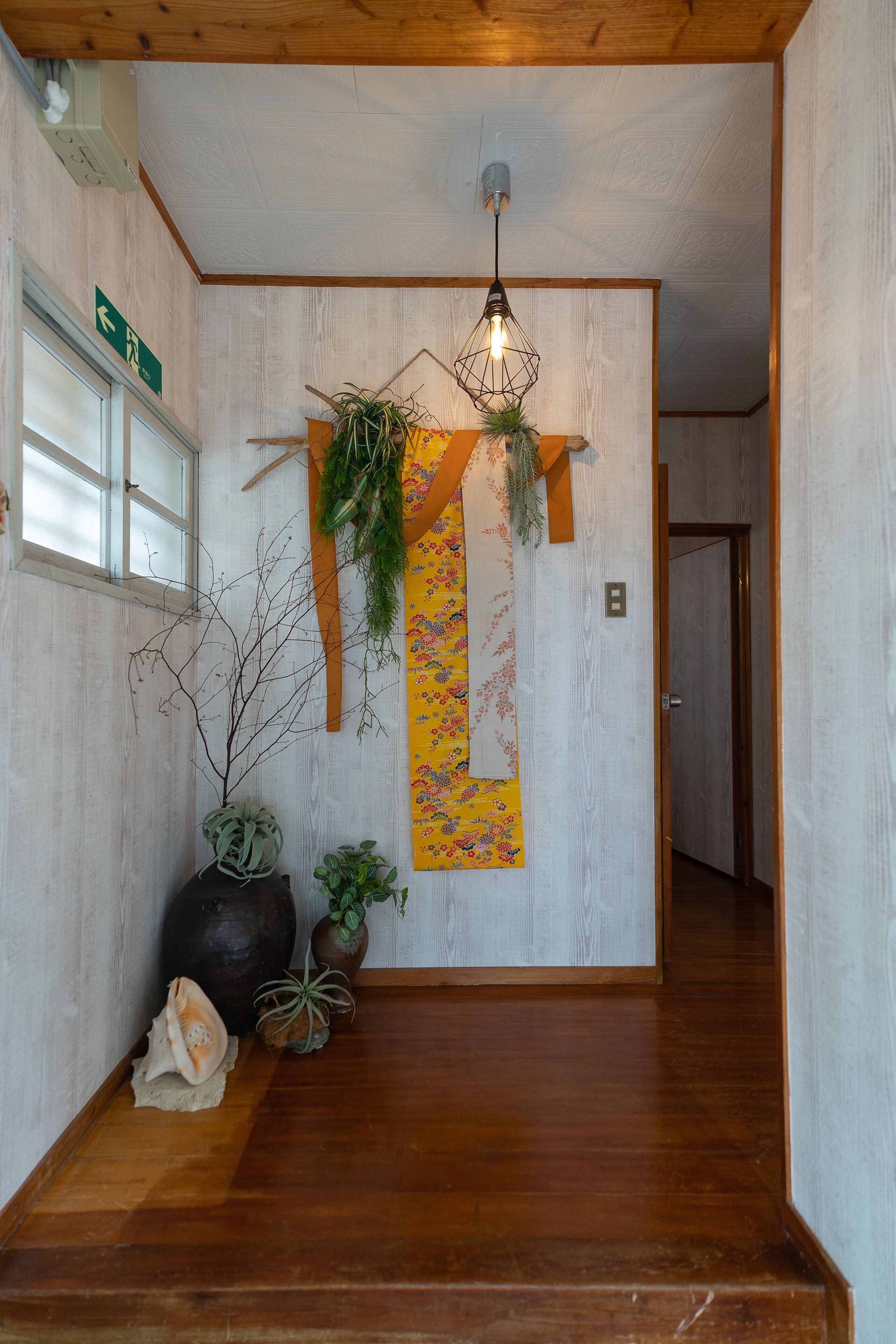 A hallway with wooden flooring, a hanging light fixture, a potted plant, a decorative wall hanging, and a door at the end.