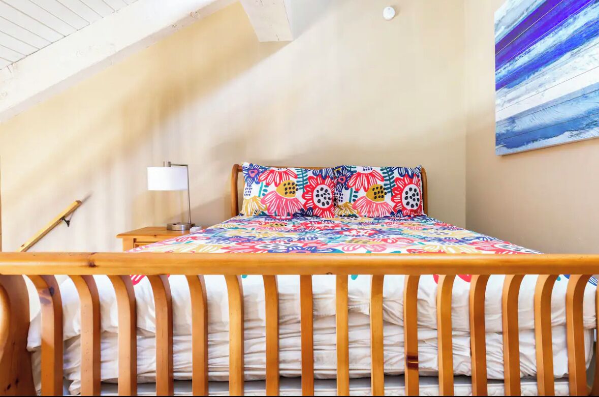 A bedroom with a wooden bed, colorful floral bedding, a bedside lamp, and a painting on the wall.