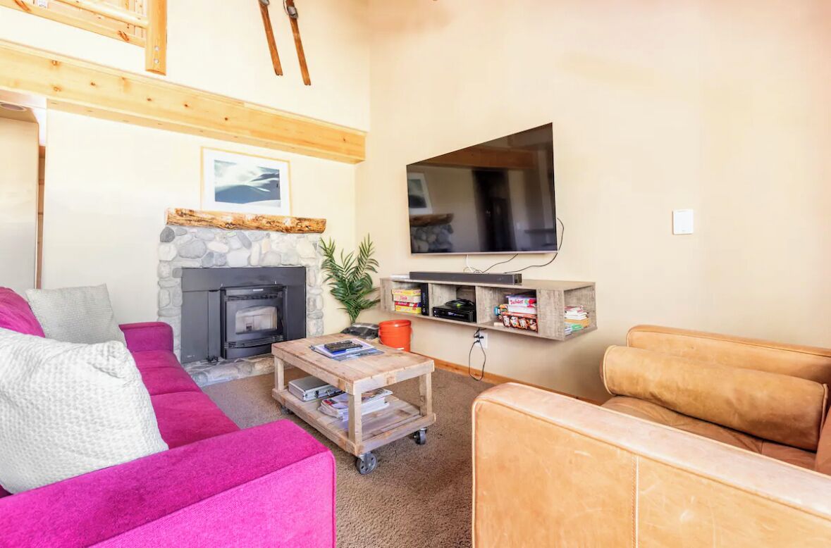 Ein gemütliches Wohnzimmer mit Kamin aus Stein, einem an der Wand befestigten Fernseher, einem Holztisch und einem rosa Sofa.