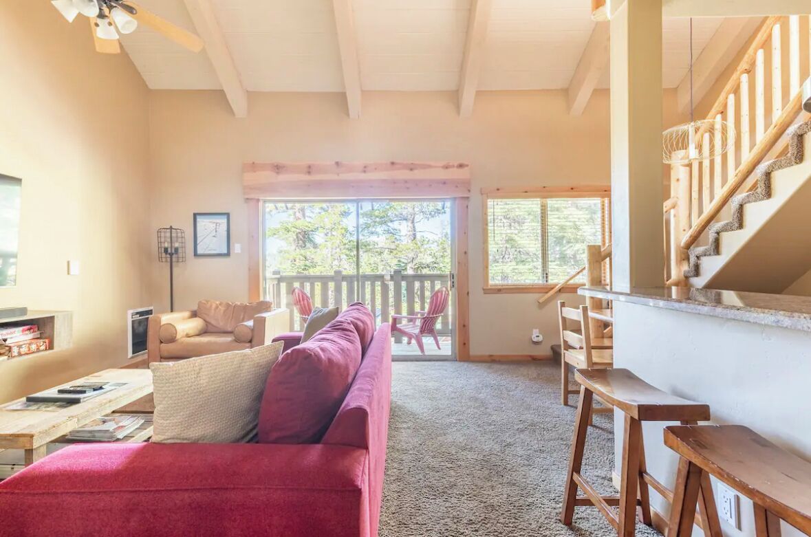 A cozy living room with a red sofa, wooden staircase, and a large window offering a view of the outdoors.