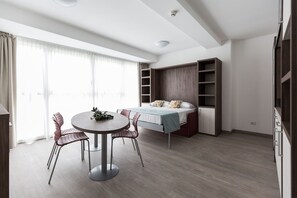 A modern bedroom with a bed, built-in shelving, a dining area with chairs, and a large window with curtains.