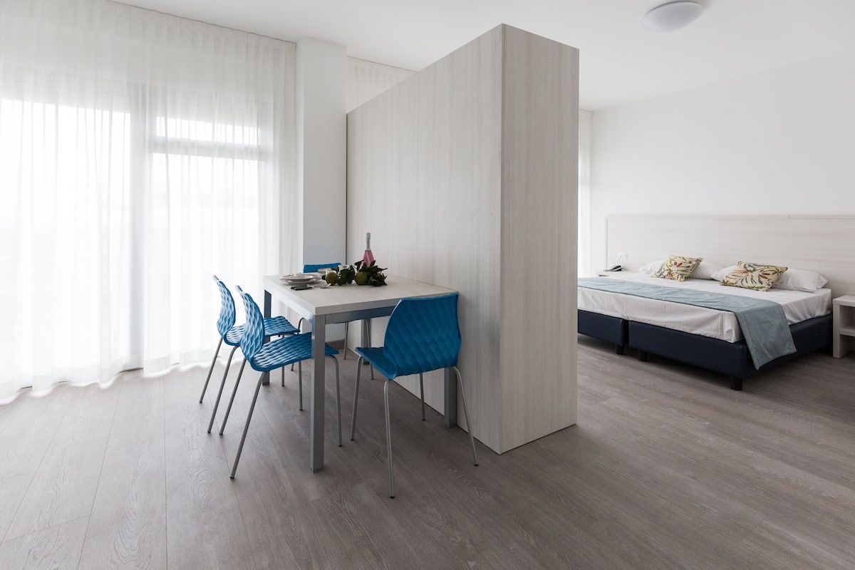 Una camera moderna con un tavolo da pranzo, sedie blu, un letto con testiera bianca e un'ampia finestra con tende leggere.