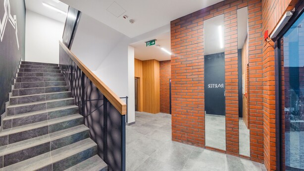 Un escalier avec une rampe en bois et des marches grises, menant à une pièce dont un mur est en briques et sur la porte est inscrit le mot « PAPETERIE ».