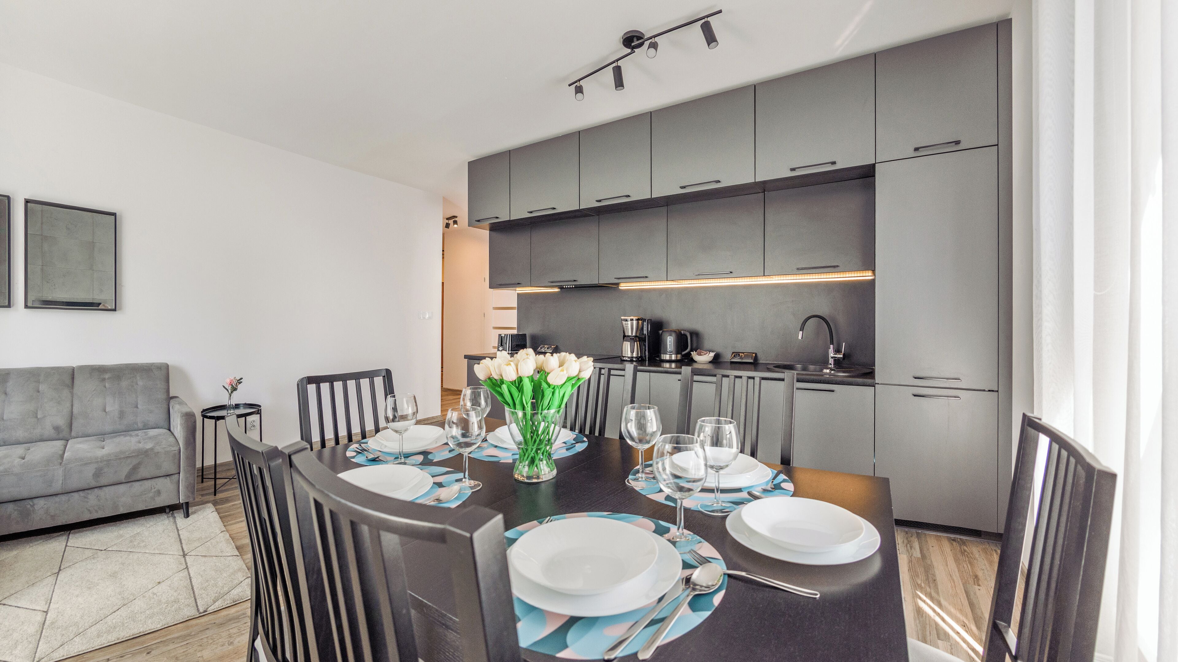 A modern dining area with a dark wooden table set for four, surrounded by grey cabinets and a kitchen in the background.