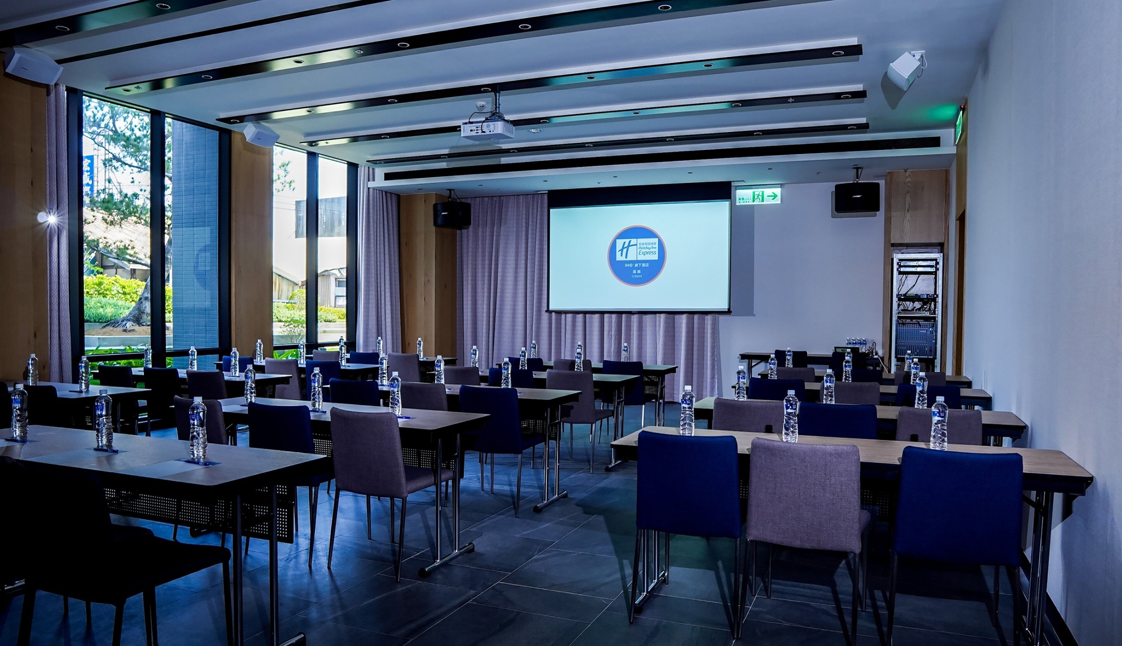 A conference room with a large screen, multiple tables, chairs, and water bottles.