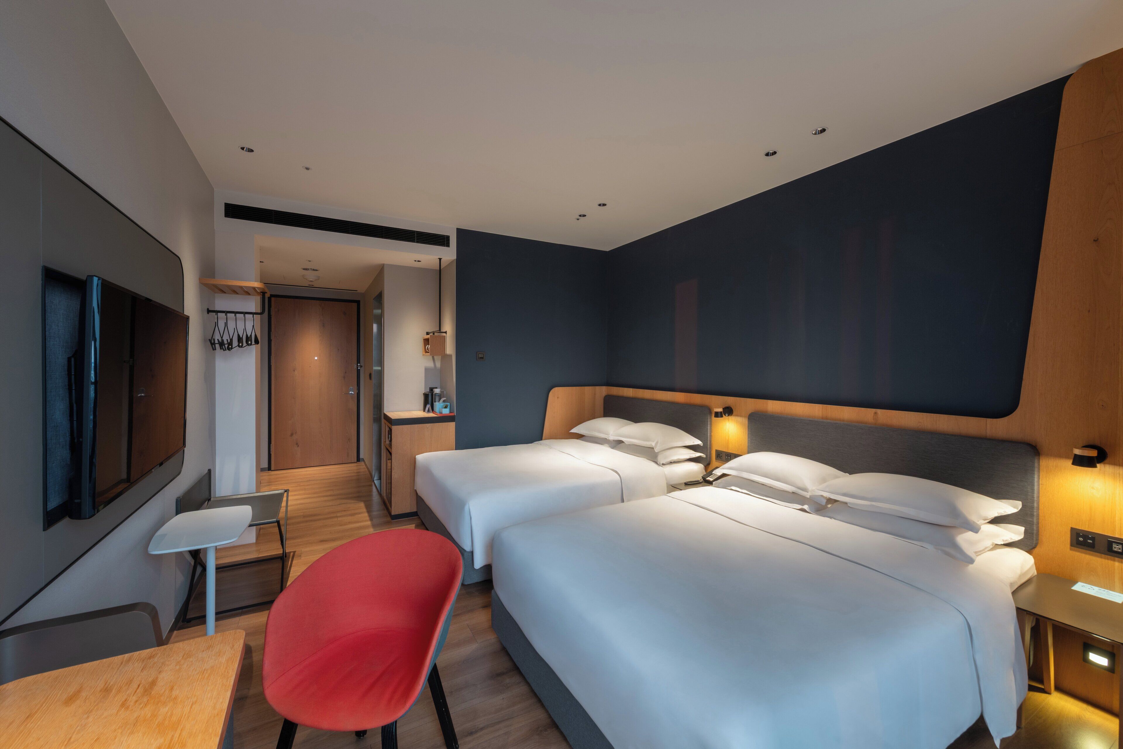 A modern hotel room with two beds, a desk, a red chair, a TV, and a wooden headboard.