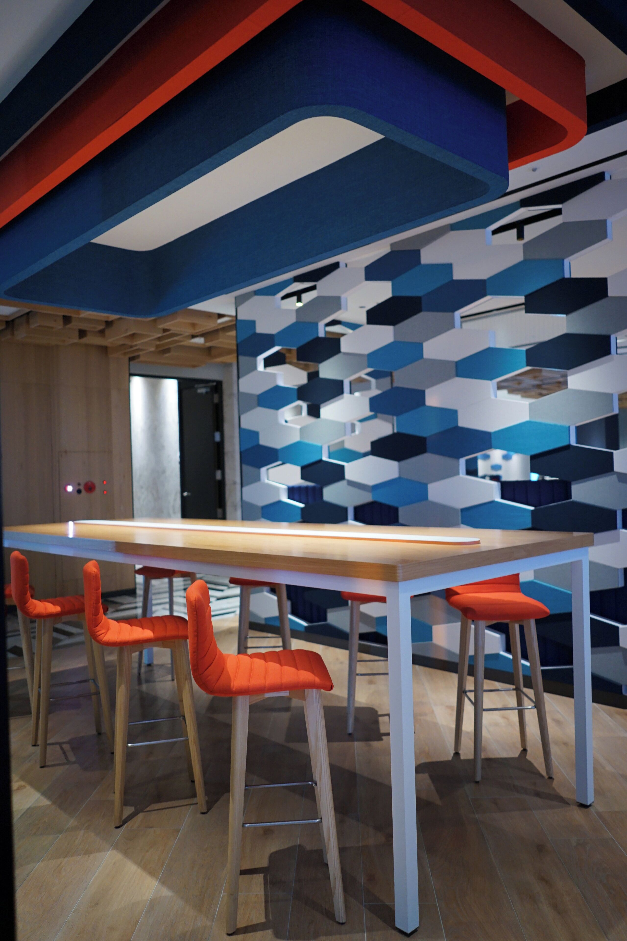 A modern interior with a long table and four orange chairs, wooden flooring, and a geometric wall design.