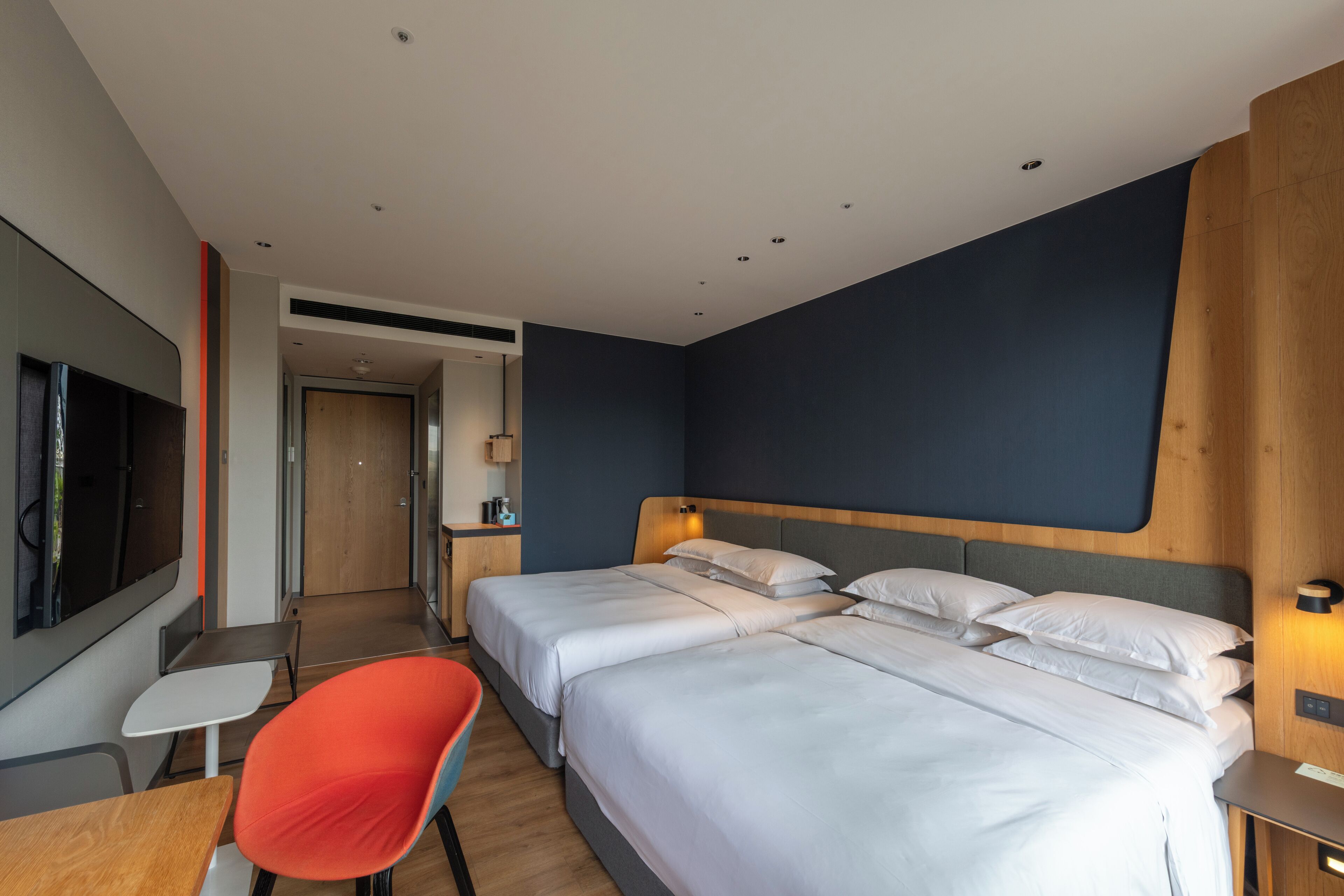 A modern hotel room with two beds, a TV, a desk, and a red chair.