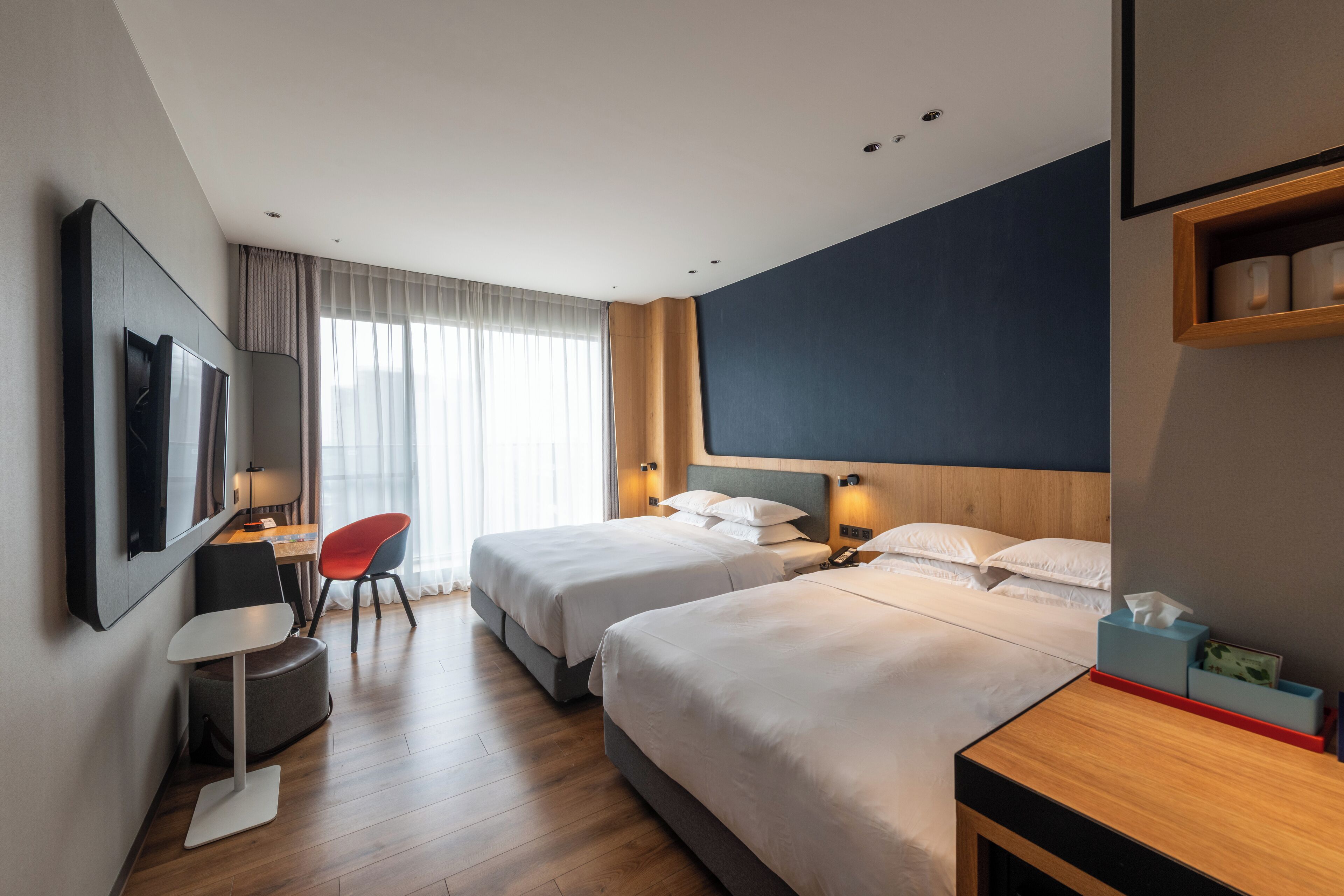 A hotel room with two beds, a desk, a chair, a TV, and a window with curtains.