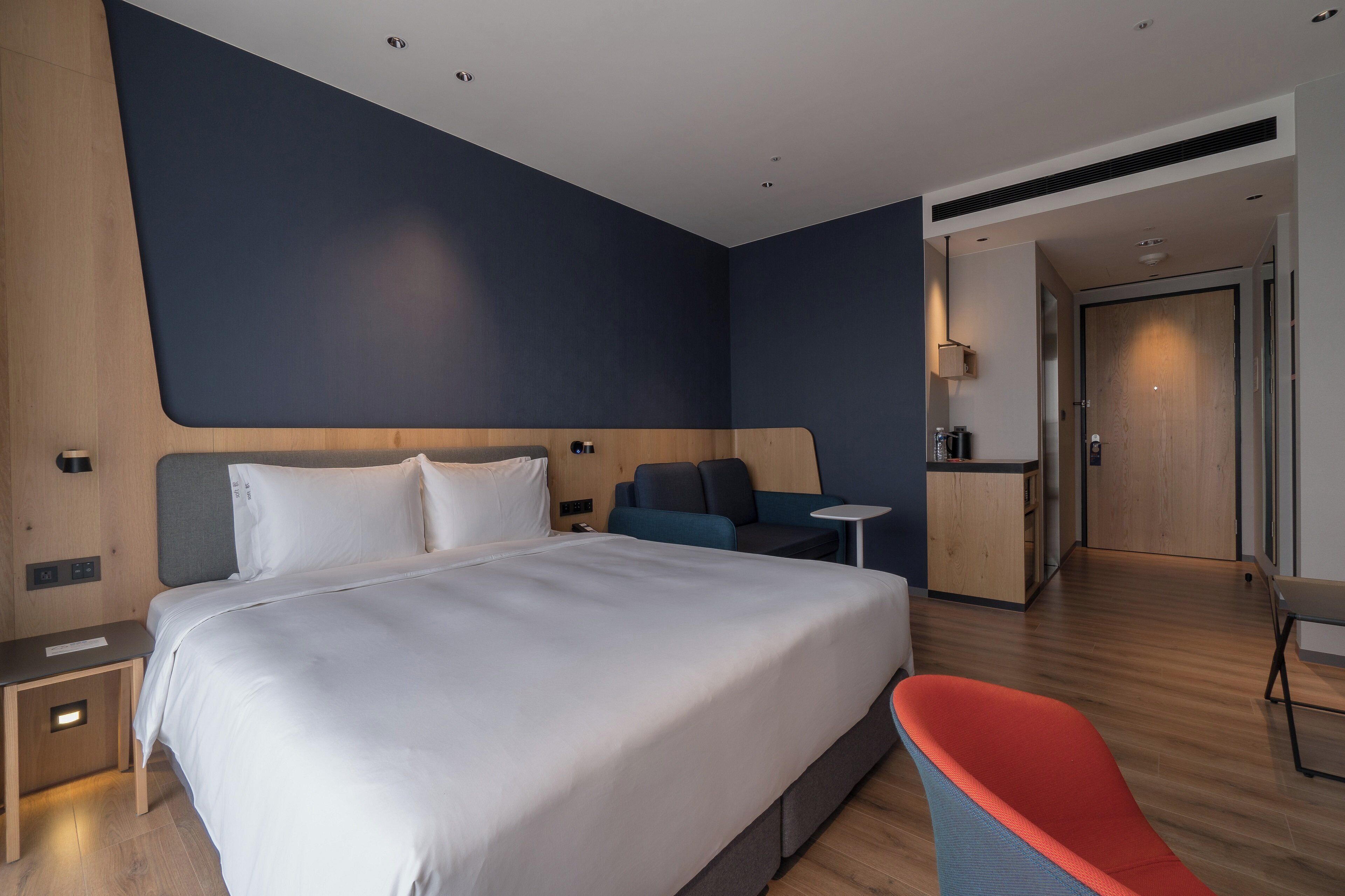 A modern hotel room with a large bed, a seating area with a small table, and a separate kitchenette.