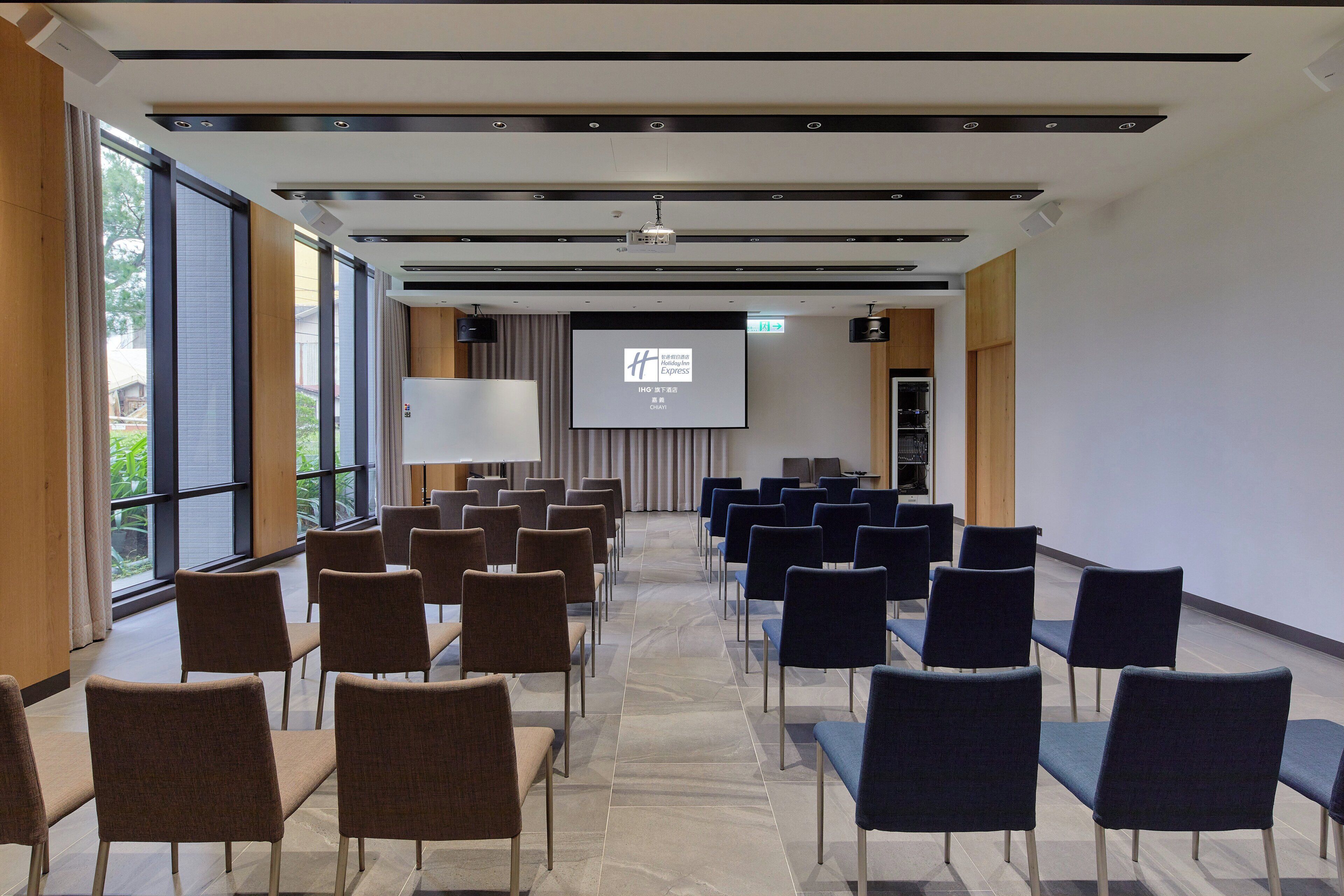 Une salle de conférence avec des rangées de chaises, un écran de projection et de grandes fenêtres.