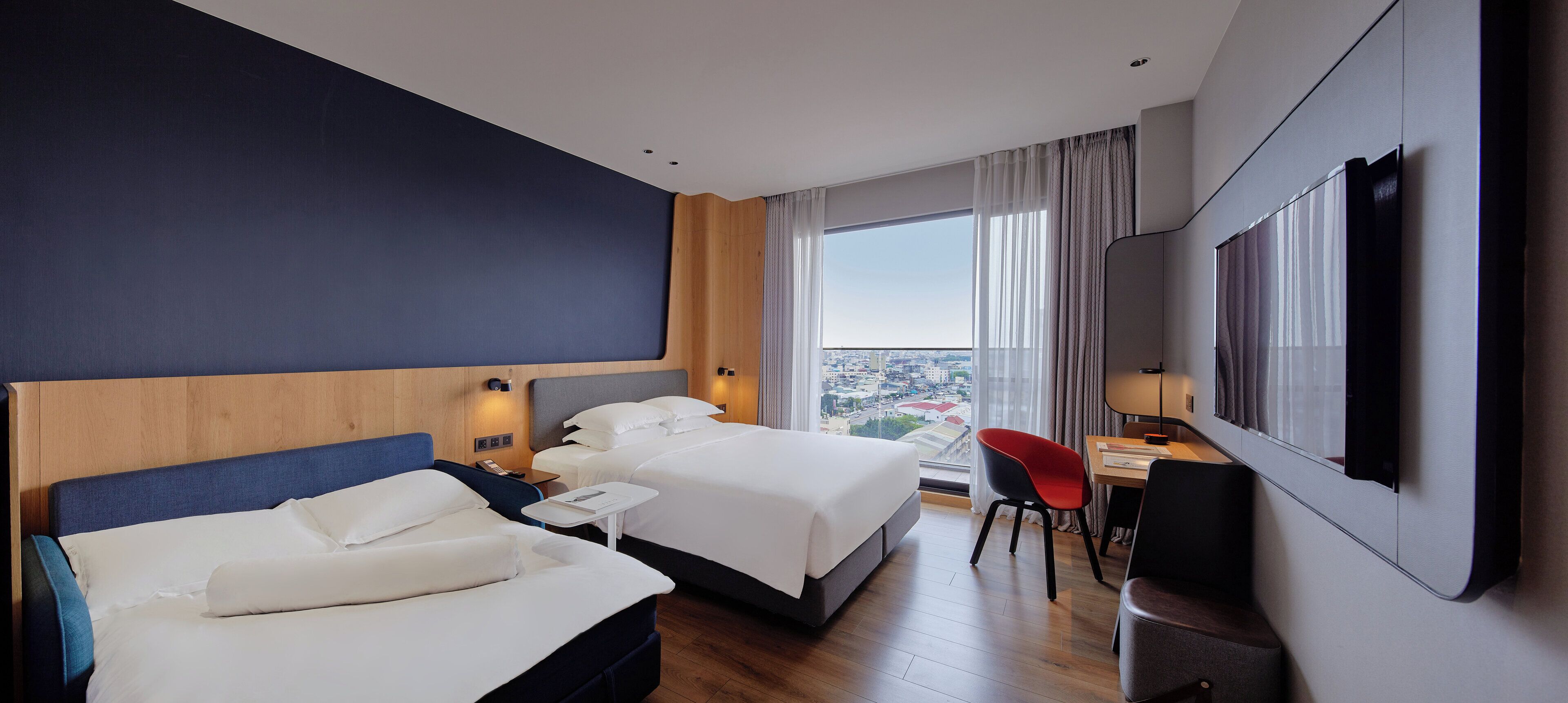 A hotel room with two beds, a desk, a chair, and a large window offering a city view.