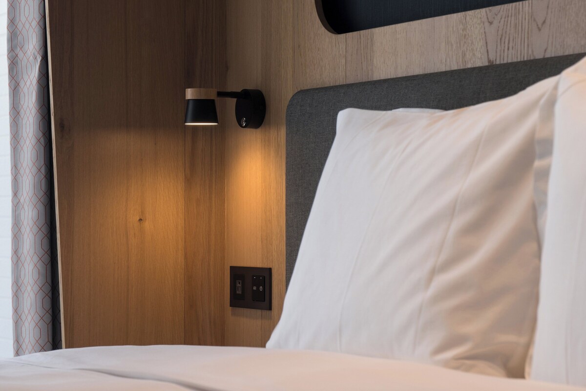 Un lit avec un oreiller blanc et une tête de lit adossée à un mur en bois, sur lequel est fixée une applique murale.