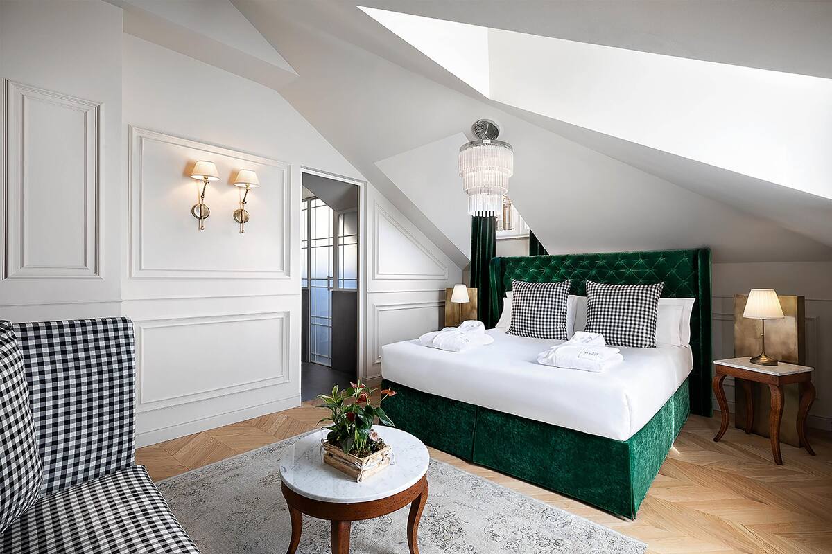 A hotel room with a checkered sofa, a bed with a green headboard, a chandelier, and a view of the sky through the sloped ceiling.