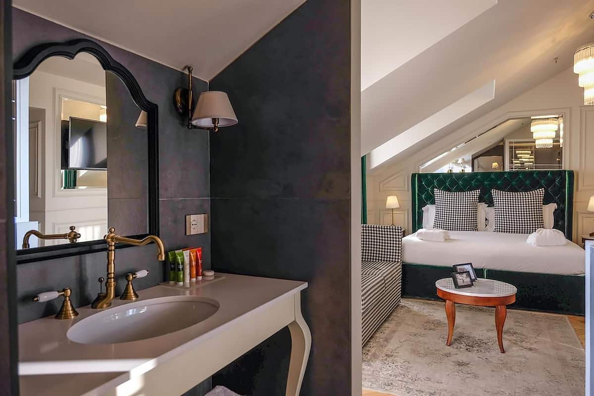 A bathroom with a large mirror, a white sink, and a dark wall. The room has a bed with a green upholstered headboard and a round wooden side table.