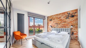 A modern bedroom with a large bed, a brick accent wall, a view of a cityscape, and a bright orange chair. - Apartamenty Sun & Snow Waterlane Island (Gdansk)