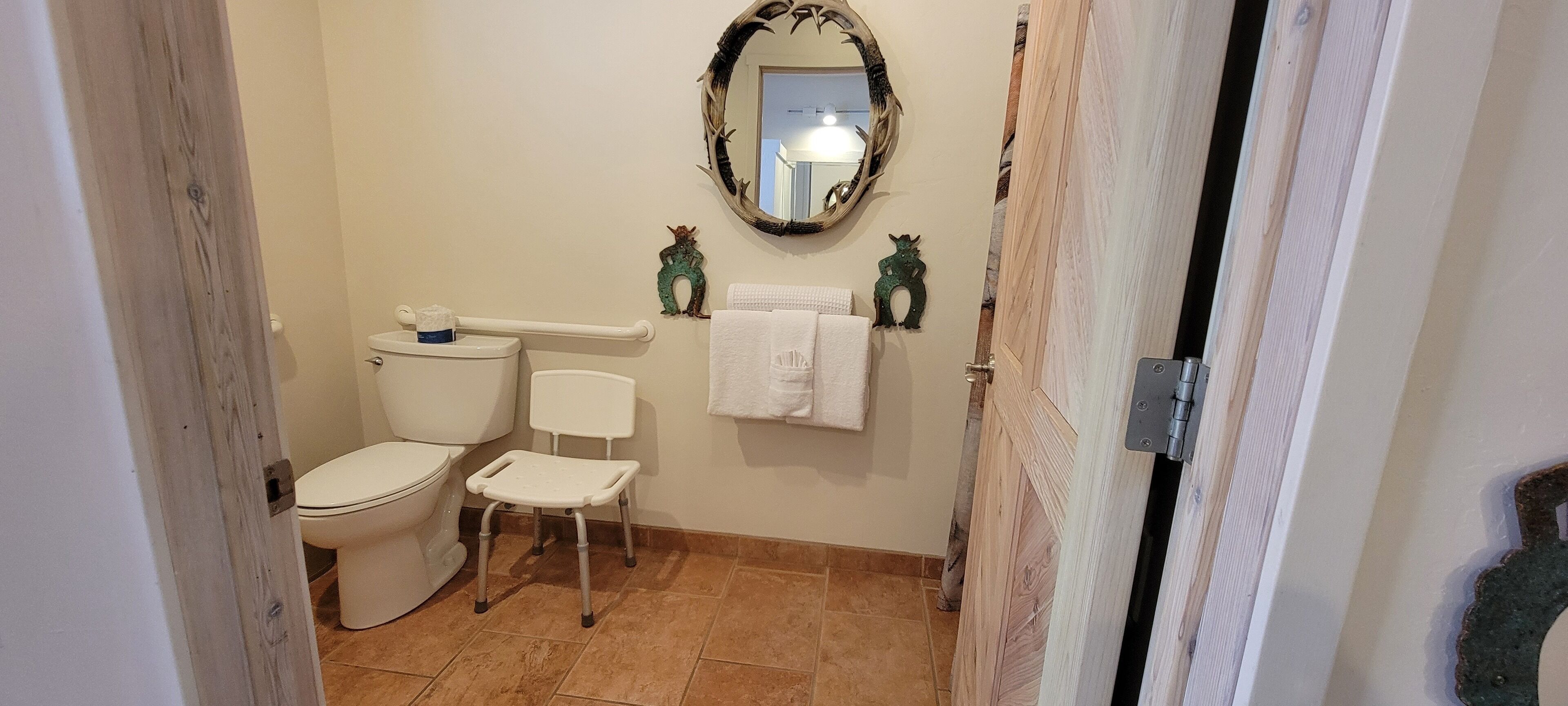 A bathroom with a toilet, a bidet, a white chair, a towel rack, and a mirror with a deer antler frame.