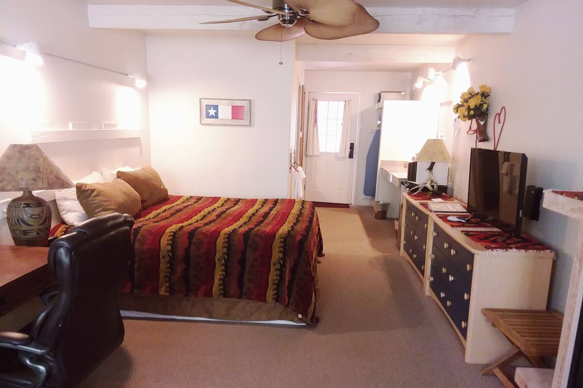 A bedroom with a bed, a desk, a chair, a dresser, a ceiling fan, and a wall with a flag and a vase with flowers.