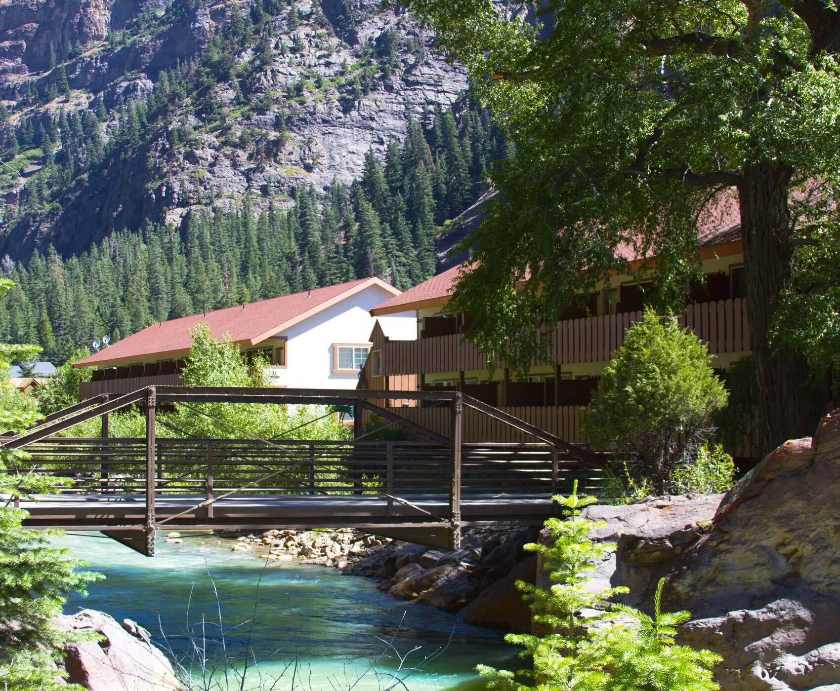 A wooden bridge over a clear river, a building with a red roof, and a mountainous backdrop.