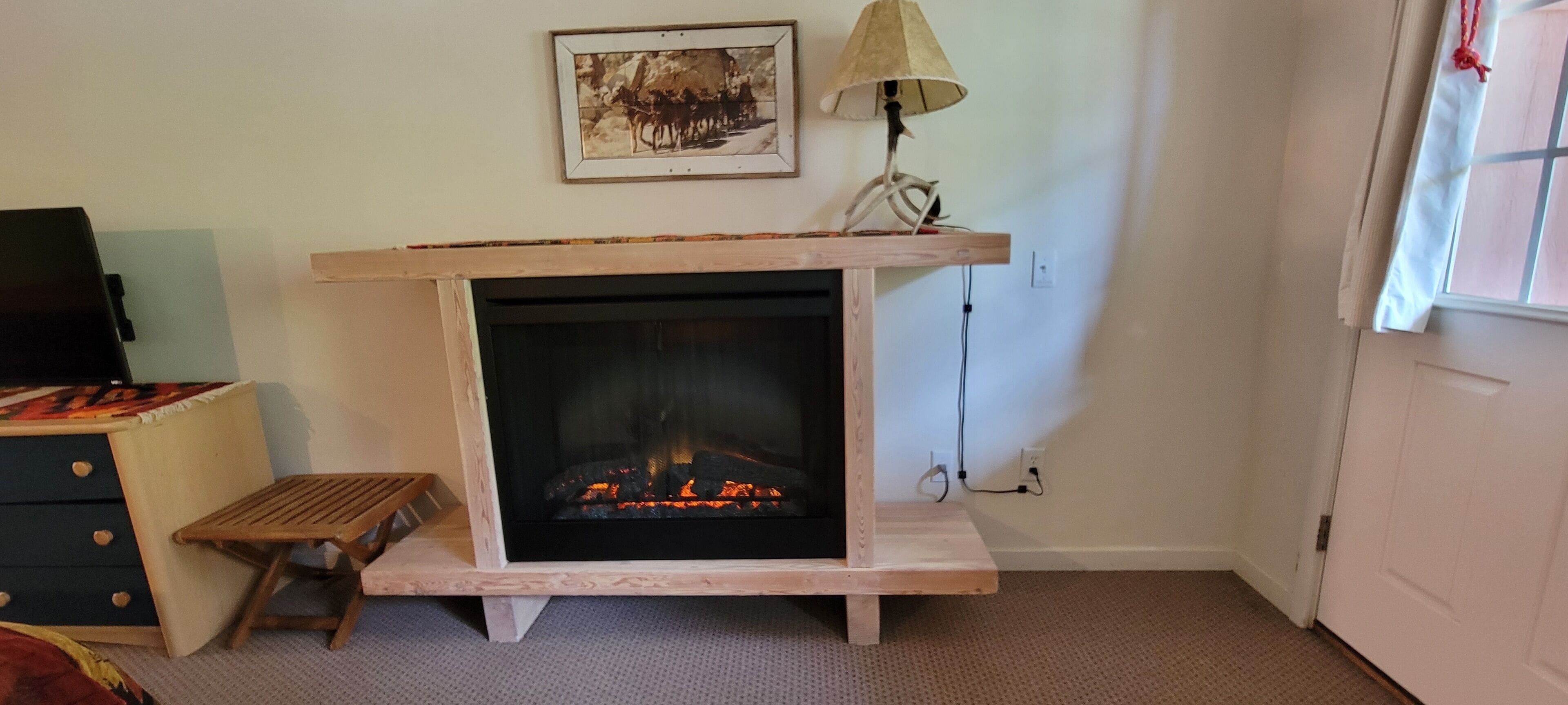 A room with a fireplace, a small wooden stool, a lamp, and a framed picture on the wall.