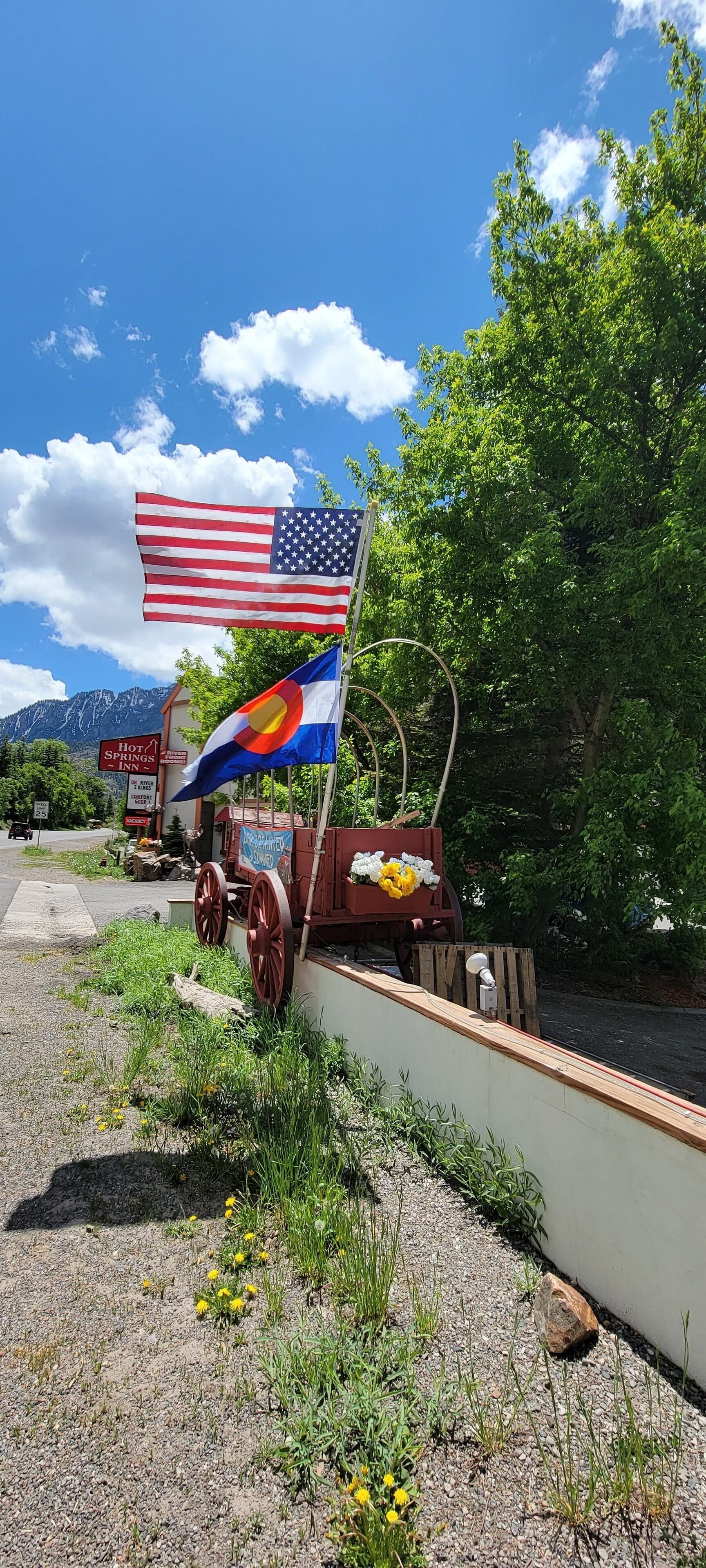 A rustic wagon with a flag, a sign for a hot springs inn, and a mountainous backdrop.