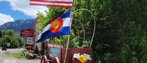 Un carruaje rústico con una bandera, un letrero de un hospedaje de aguas termales y un paisaje montañoso de fondo.