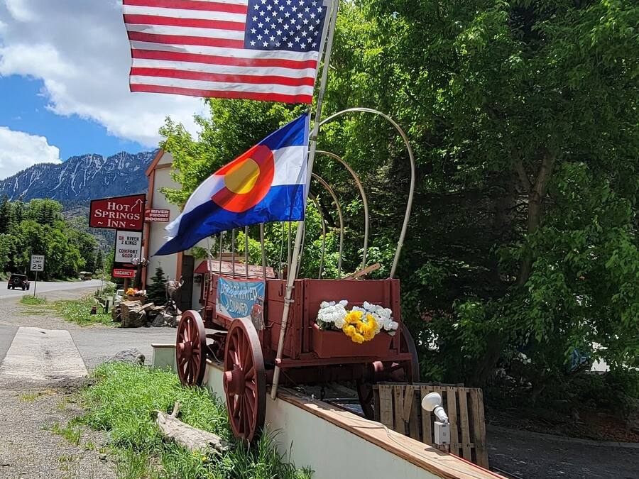 A rustic wagon with a flag, a sign for a hot springs inn, and a mountainous backdrop.