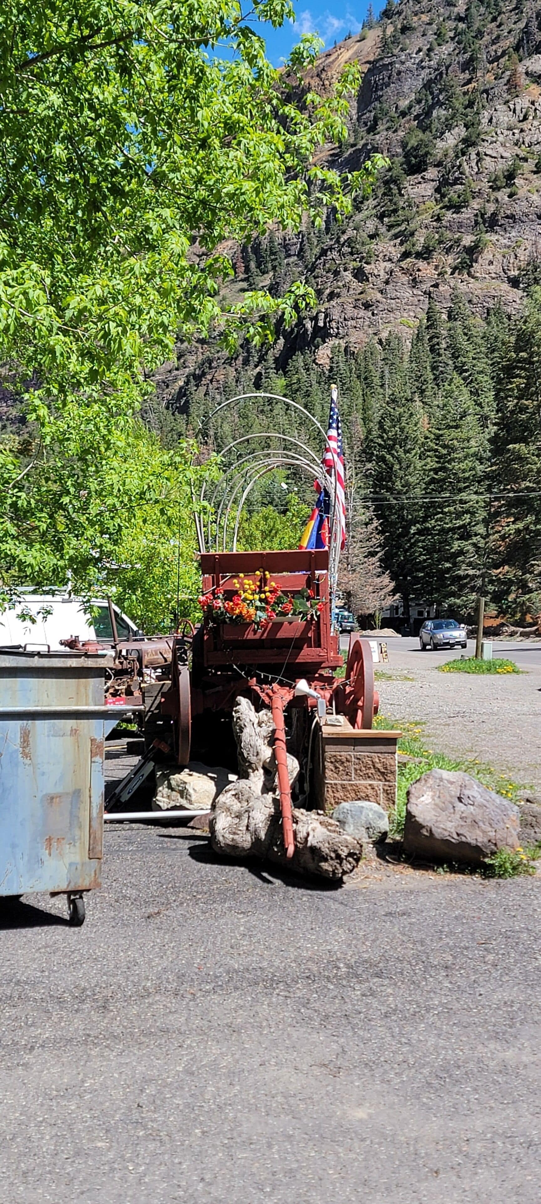 Ein alter Traktor mit amerikanischer Flagge, geparkt auf einem Kiesgrund mit Bäumen und einem Berg im Hintergrund.