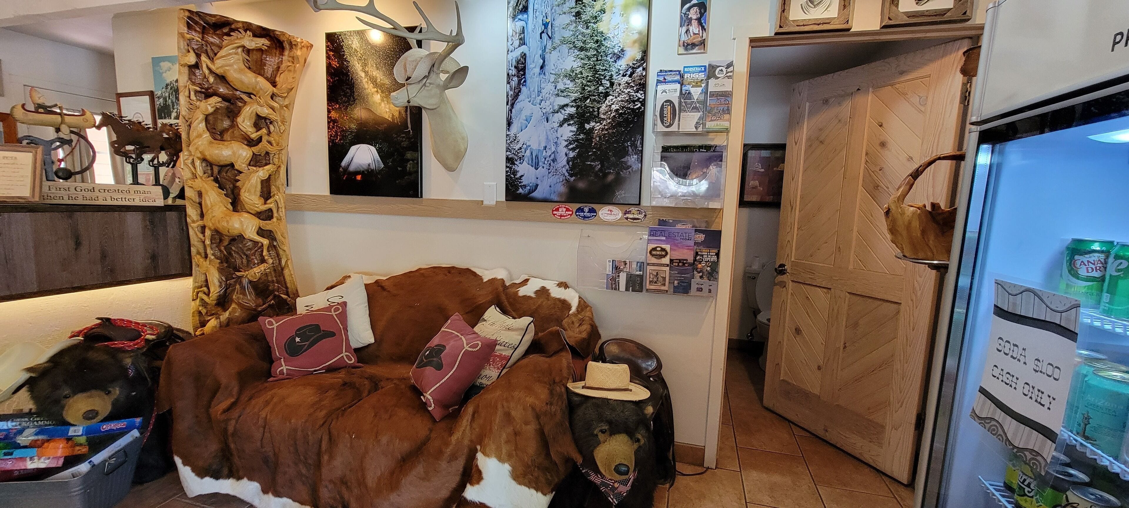 Un cuarto acogedor con un sofá con temática de vaca, un refrigerador lleno de bebidas y diversas decoraciones en las paredes, incluyendo una cabeza de ciervo y carteles.