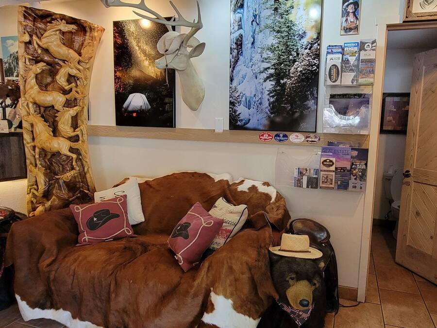 Un cuarto acogedor con un sofá con temática de vaca, un refrigerador lleno de bebidas y diversas decoraciones en las paredes, incluyendo una cabeza de ciervo y carteles.
