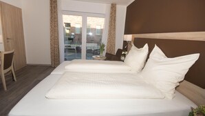 A hotel room with two beds, a wooden wardrobe, a small table with a plant, and a view of the outside through a glass door. - Hotel Sonne am Meer (Norden)