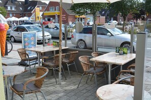 Espace extérieur aménagé avec des chaises et des tables en osier, un panneau affichant le menu et une vue sur la rue, où sont garées des voitures.